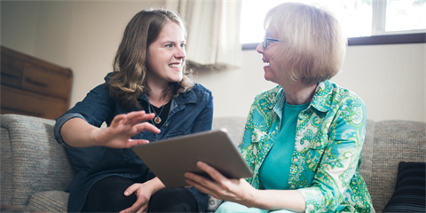 A young woman sits on a couch with an older women, instructing her how to use a tablet