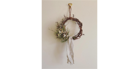 A wreath made from twisted vines and natural materials with flowers and foliage and a natural fibre material hanging from the bottom.